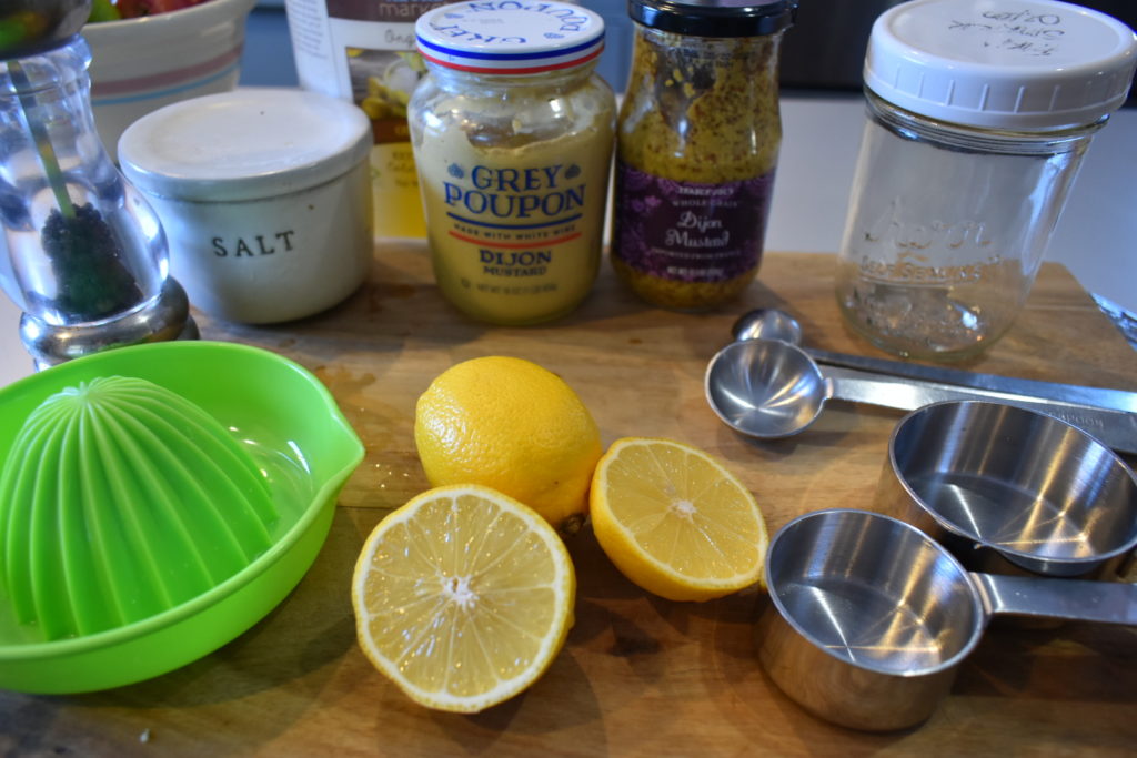Lemons, measuring cups and teaspoons, mustard, salt, pepper and a glass jar on a wooden cutting board. 